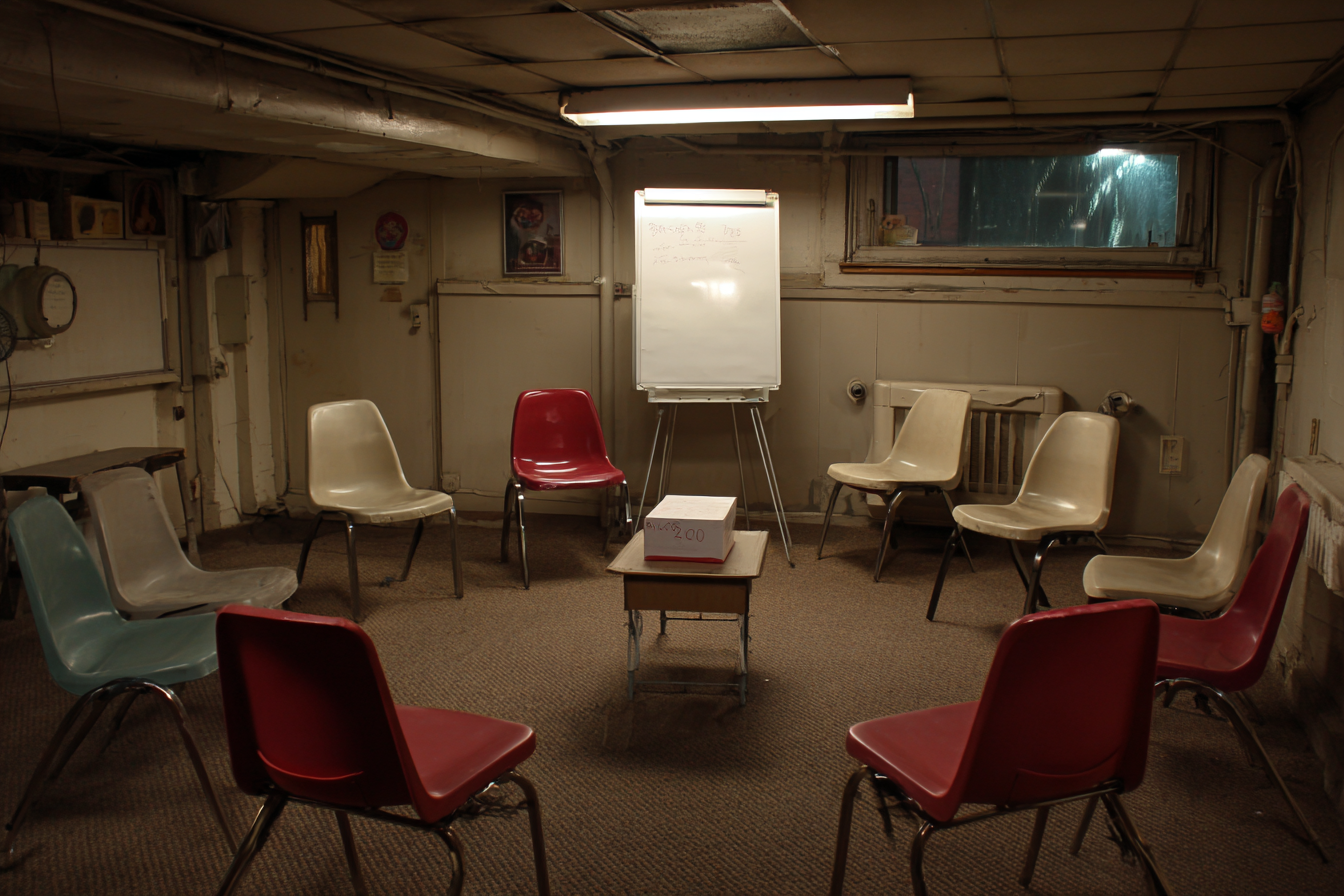 Mismatched chairs arranged in a circle in a basement room, with a flipchart easel and a small table at the center